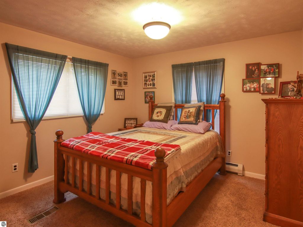 Cozy bedroom featuring a wooden bed with a plaid blanket, soft lighting, blue curtains, and framed family photos on the walls, reflecting the inviting atmosphere of the 3-bed, 2-bath home for sale in Copemish, MI.