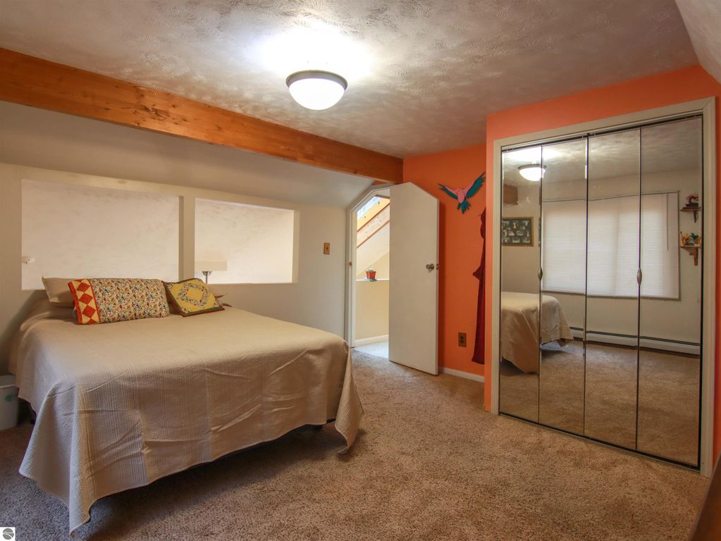Cozy bedroom in saltbox style home featuring a queen-sized bed with decorative pillows, mirrored closet doors, and warm orange walls, showcasing a comfortable living space within the 3-bedroom, 2-bath property for sale in Copemish, MI.