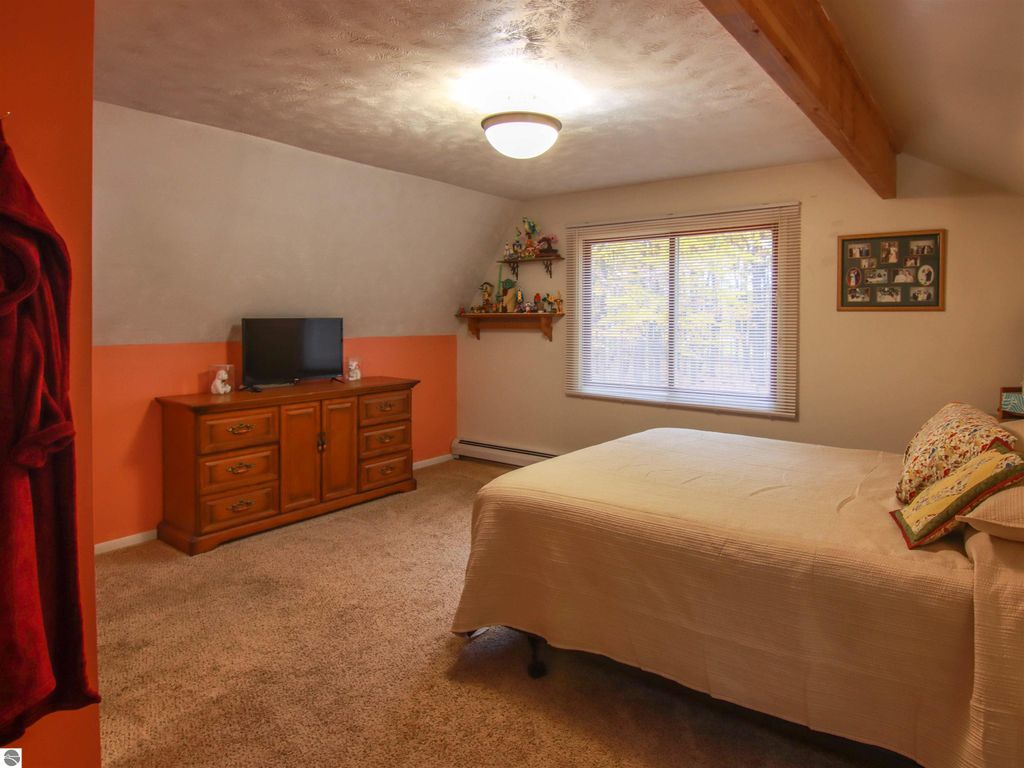 Cozy bedroom in a saltbox style home featuring a queen-sized bed, wooden dresser, and TV, with natural light from a window overlooking wooded views.