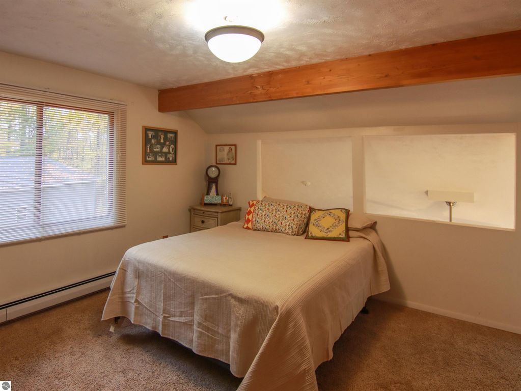 Cozy bedroom with a queen-sized bed, decorative pillows, large window with blinds, and wooden beam ceiling, showcasing a warm and inviting atmosphere in a 3-bedroom home in Copemish, MI.