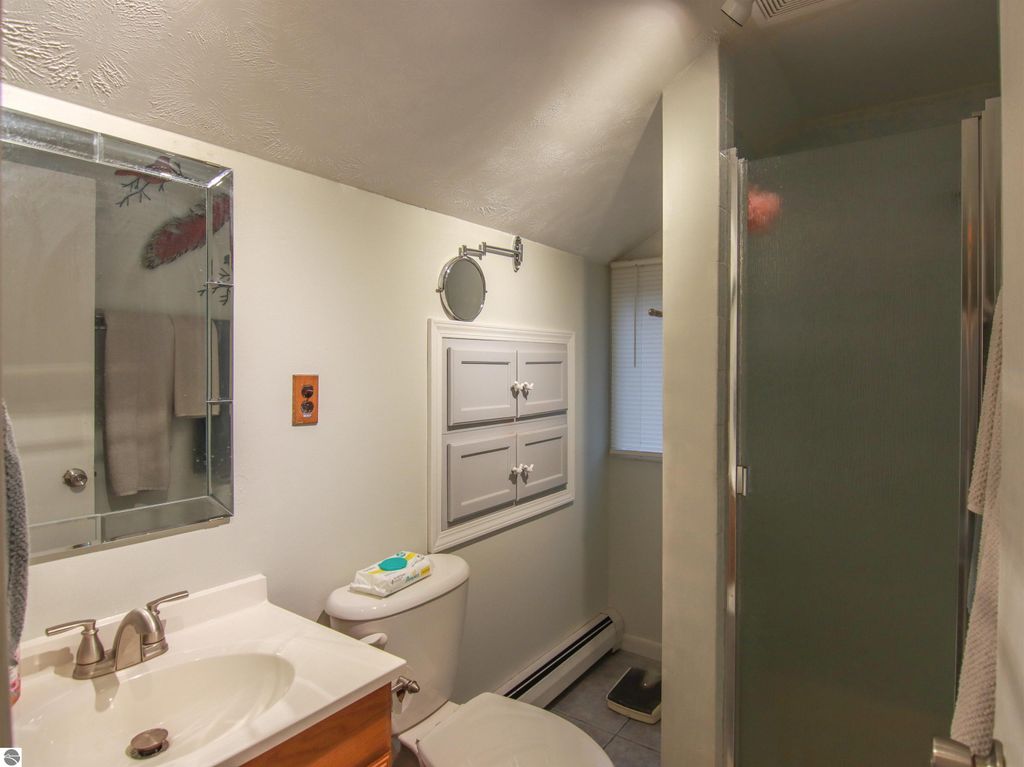 Bathroom interior featuring a sink, toilet, and shower, with light-colored walls, a mirror, and storage cabinets, part of a 3-bed, 2-bath home in Copemish, MI.