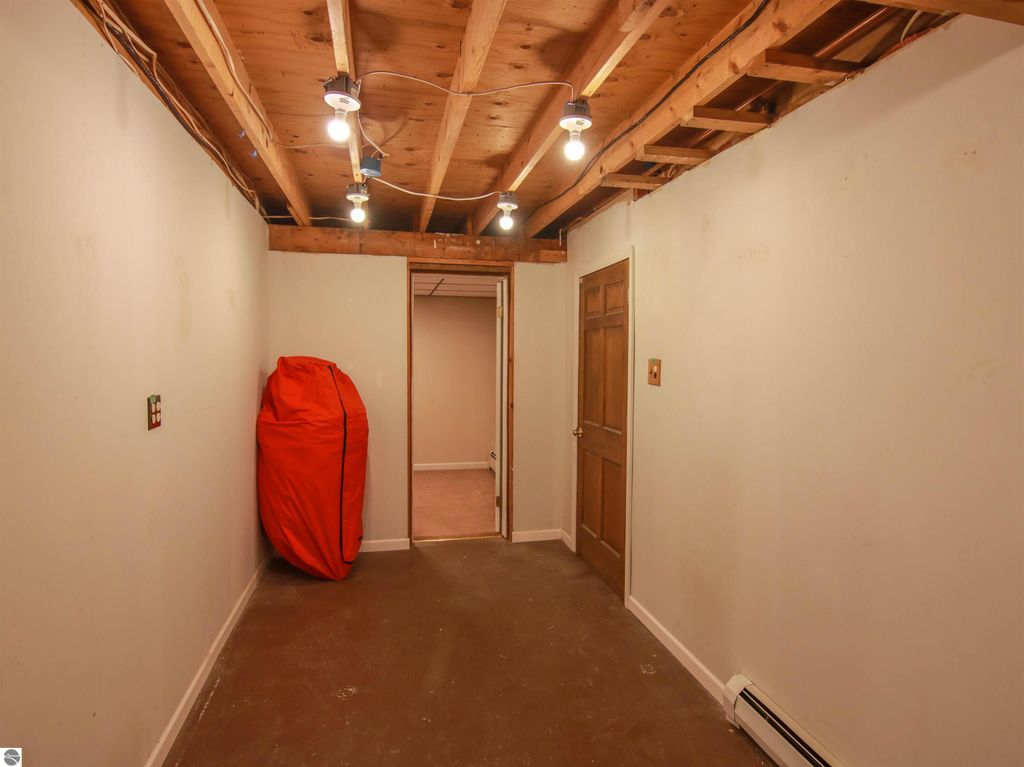 Lower level storage room with exposed wooden beams, overhead lighting, and an orange storage bag against the wall, leading to a doorway.