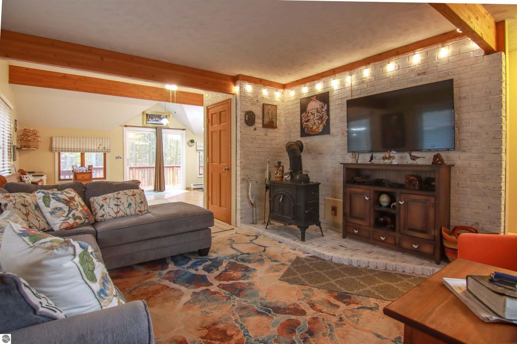 Cozy living room with a woodstove, plush gray sofa, colorful area rug, and a TV mounted on a brick wall, featuring large windows that provide views of the wooded surroundings, highlighting the inviting atmosphere of the 3-bedroom, 2-bath home on 8.75 acres in Copemish, MI.