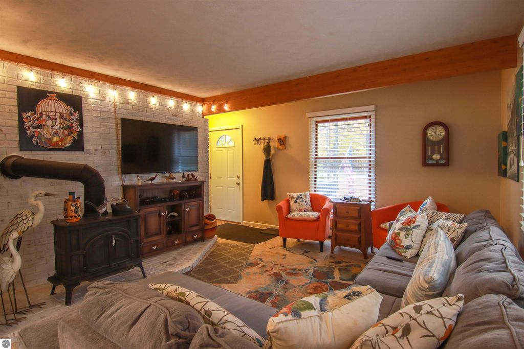 Cozy living room in a saltbox style home featuring a woodstove, plush sofa with decorative pillows, and bright orange accent chairs, complemented by wooden beams and natural light.