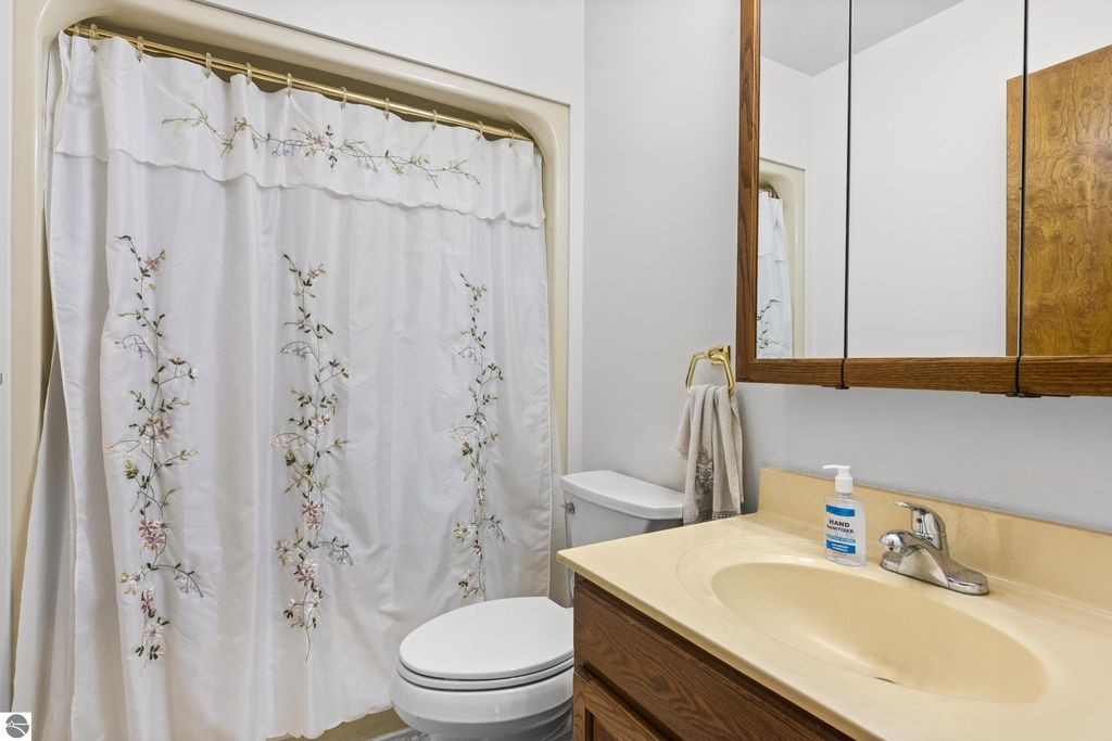 Bathroom featuring a white shower curtain with floral embroidery, beige countertop with sink, mirror, and wooden cabinetry, emphasizing cleanliness and functionality in the property at 396 Davenport Lane, Cadillac, MI.