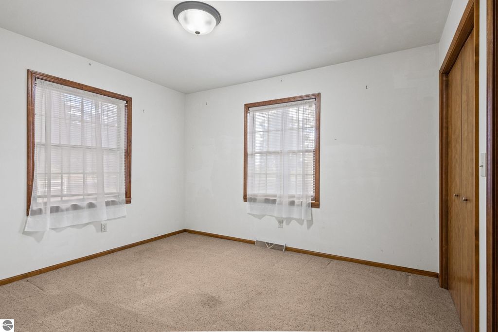 Empty bedroom with two windows featuring sheer curtains, beige carpet, and a wooden door, showcasing a bright and airy atmosphere in the Cadillac, MI home listing.