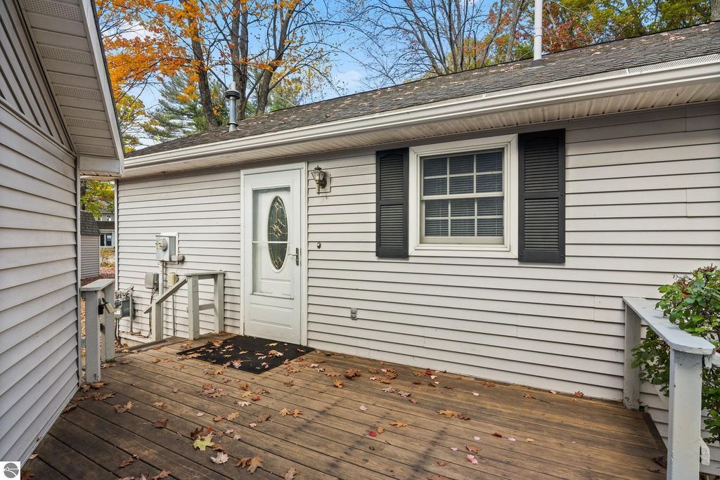 Exterior view of the entrance to a home at 396 Davenport Lane, Cadillac, MI, featuring a white door with decorative glass, black shutters, and a wooden deck surrounded by autumn leaves.