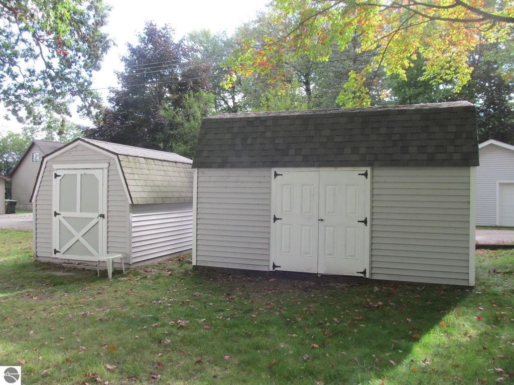 Two storage sheds in a wooded backyard setting, featuring a larger shed with double doors and a smaller shed with a single door, surrounded by green grass and trees, relevant to the property listing at 396 Davenport Lane, Cadillac, MI.
