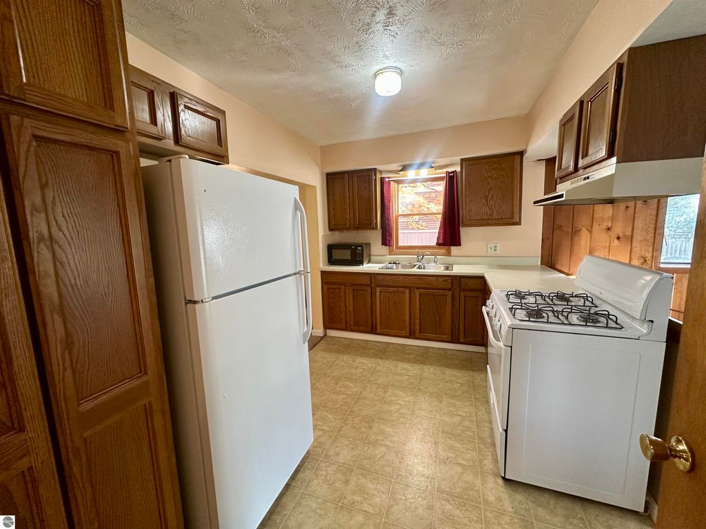 Kitchen interior featuring white refrigerator, gas stove, and wooden cabinets, with a window and sink illuminated by natural light, showcasing a clean and updated space in Traverse City home listing.