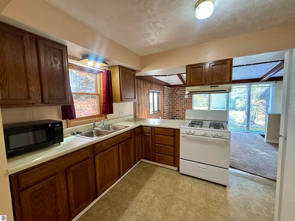 Kitchen interior featuring wooden cabinets, gas stove, double sink, and microwave, with a view of a sunlit living area through large windows, showcasing a cozy and updated space in a Traverse City home listing.