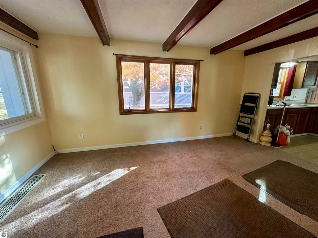 Bright interior of a residential living room featuring large window with views of foliage, exposed wooden beams, and carpeted floor, showcasing the space's potential for various layouts in the Traverse City home listing.