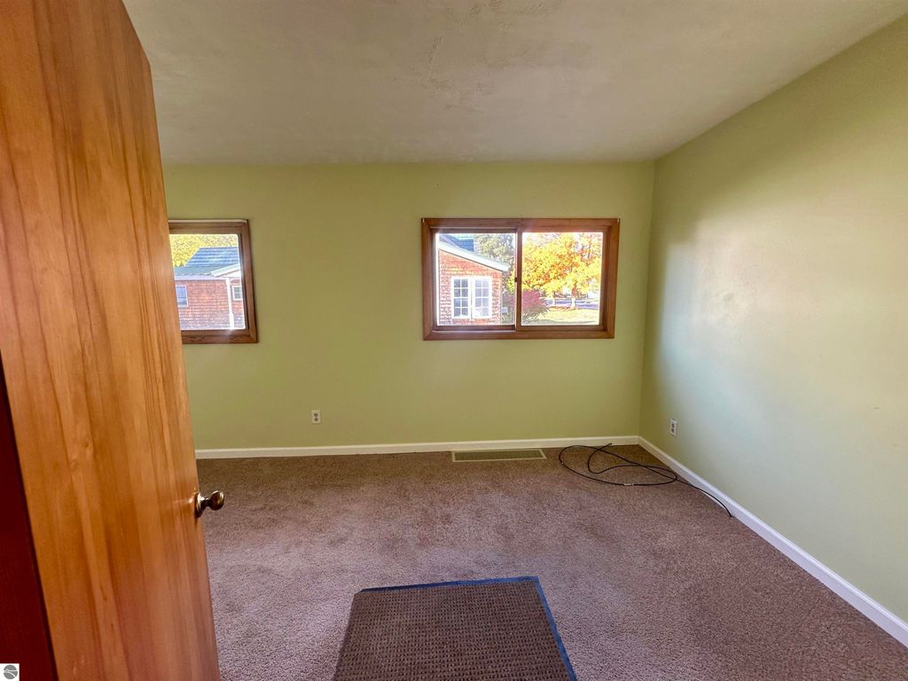 Interior view of a light green room with two windows, showcasing a cozy space suitable for a bedroom, featuring carpet flooring and a wooden door, highlighting the home's charm in Traverse City, MI.