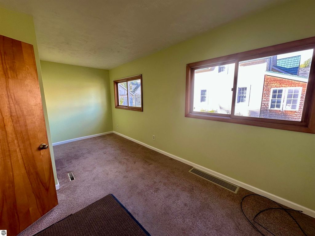 Interior view of a light green bedroom with windows, showcasing carpeted flooring and a wooden door, reflecting the cozy atmosphere of the Traverse City home for sale at 209 W Fifteenth Street.