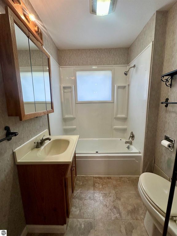 Bathroom with a shower-tub combination, sink with wooden cabinet, wall-mounted mirror, and beige tile flooring, showcasing a clean and bright interior.