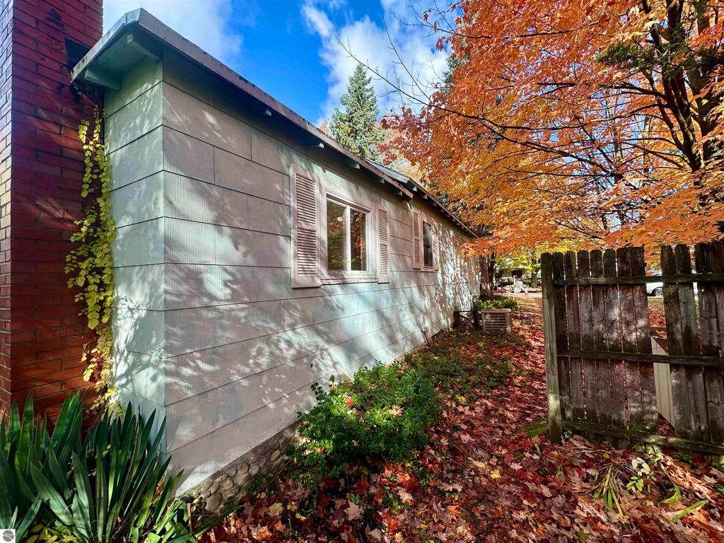 Side view of a single-story house with light blue siding, white shutters, and a brick chimney, surrounded by autumn foliage and a wooden fence, located at 209 W Fifteenth Street, Traverse City, MI.