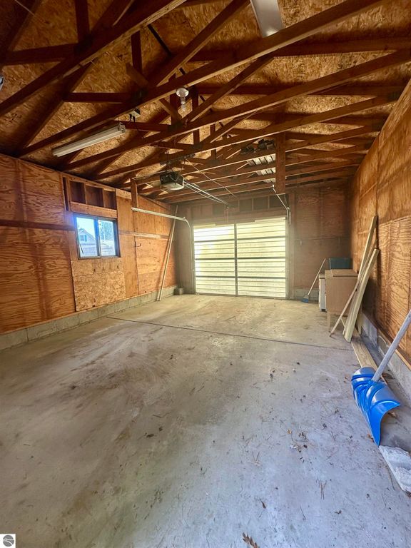 Interior of a garage with exposed wooden beams, concrete floor, and a garage door partially open, featuring a blue snow shovel and various tools against the wall, highlighting storage space for the property at 209 W Fifteenth Street, Traverse City, MI.
