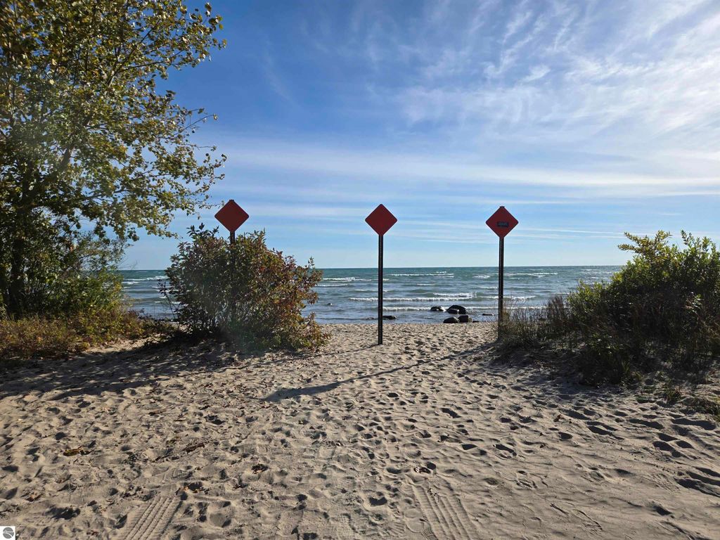 Beach access path leading to Lake Huron, flanked by three warning signs, surrounded by sandy terrain and vegetation, with a clear sky above.