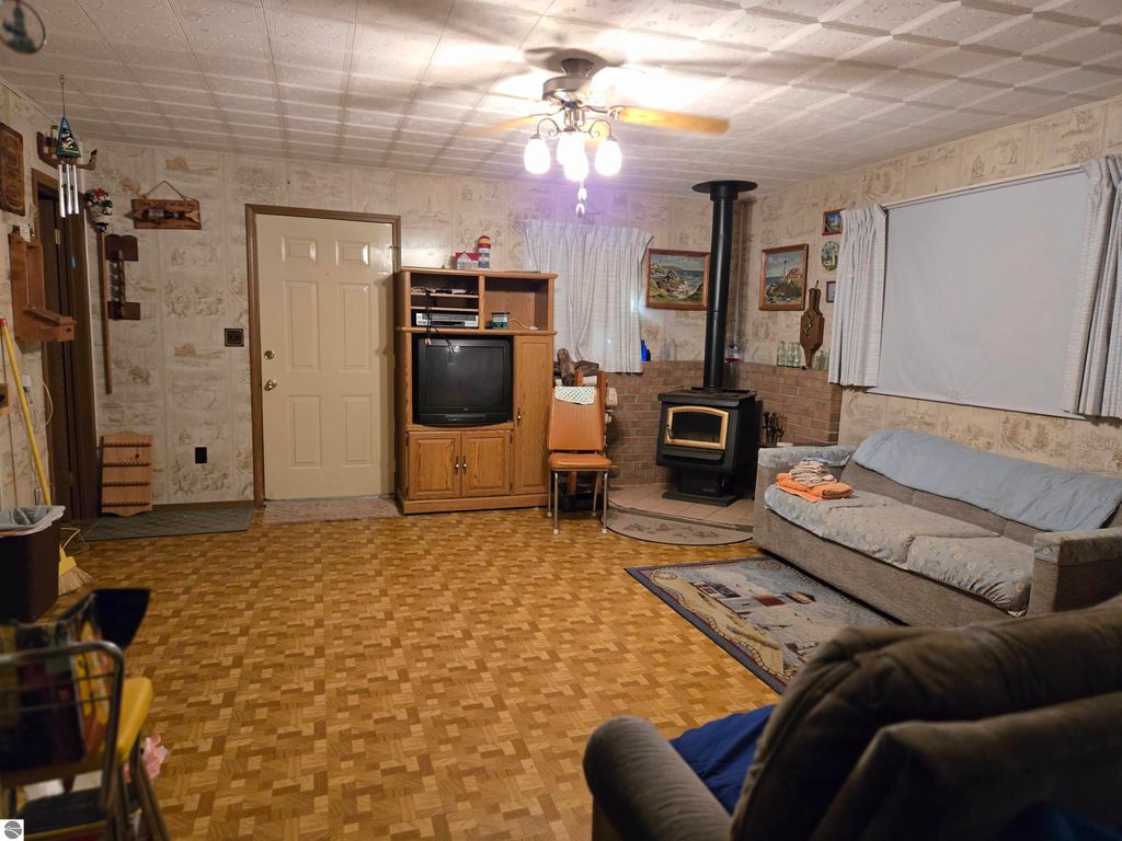 Cozy living room interior featuring a gray sofa, wooden furniture, a television, and a wood stove, set in a rustic style with a patterned floor, ideal for a 2-bedroom home in Harrisville, MI.