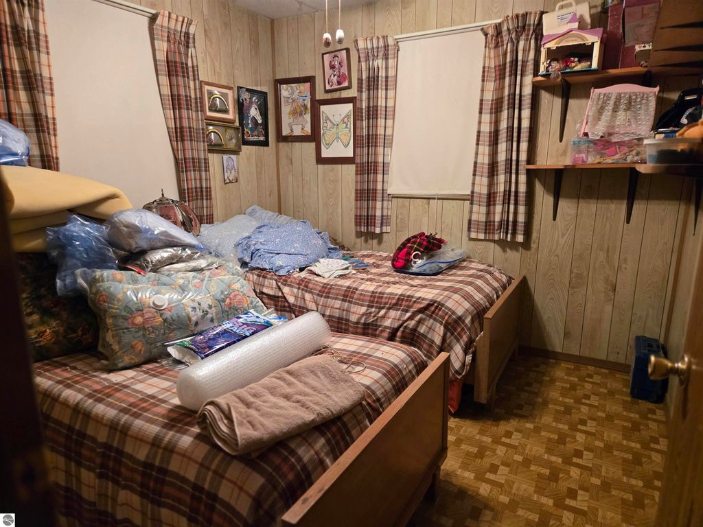 Cozy bedroom with two beds, plaid bedding, and cluttered decor, featuring patterned curtains and wooden paneling, showcasing potential for a residential retreat in Harrisville, MI.