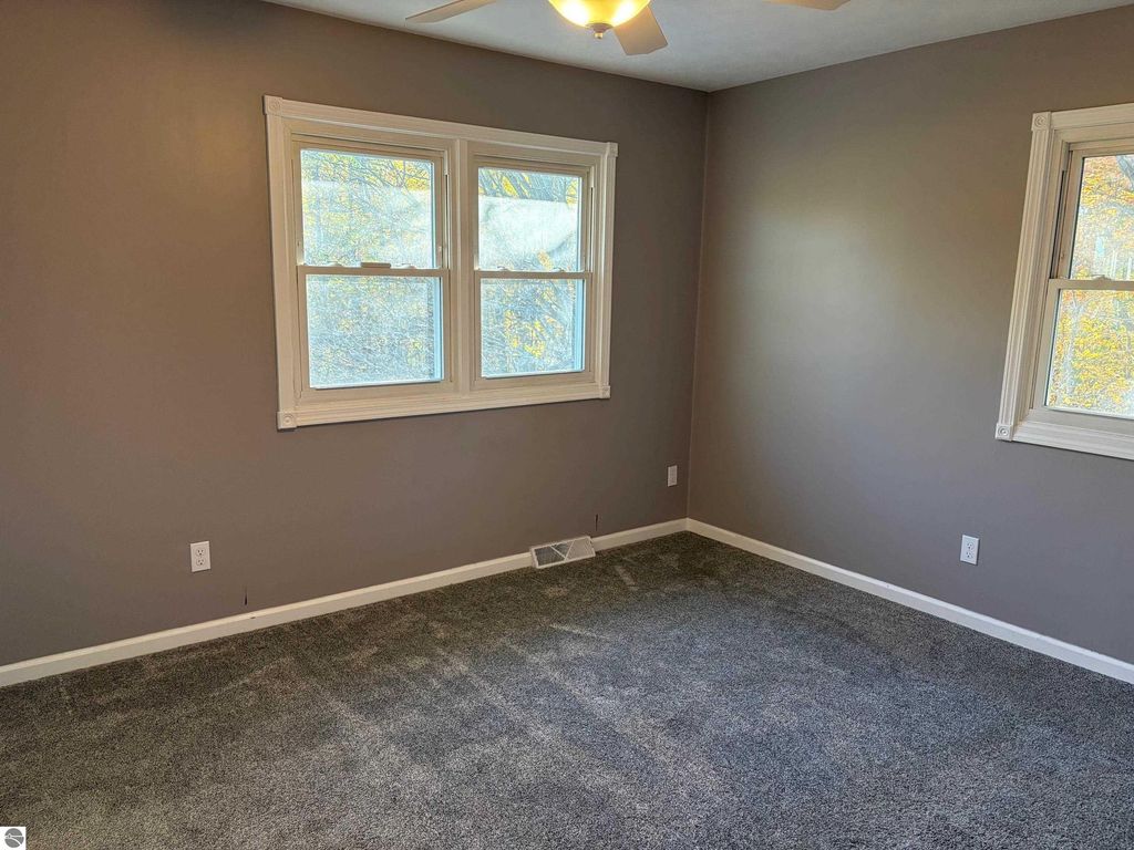 Interior view of a vacant room featuring gray walls, large windows allowing natural light, and new carpet, part of the ranch-style home located at 8721 Glover Lake Road, Bear Lake, MI.