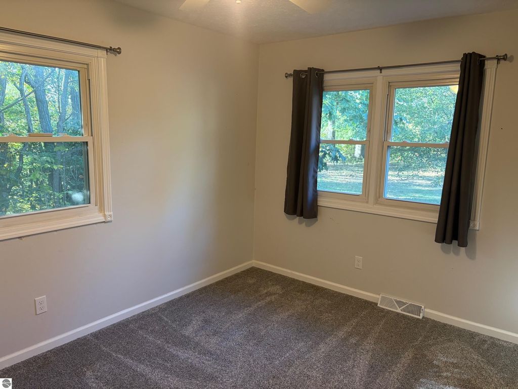 Interior view of a bedroom featuring two windows with curtains, carpeted flooring, and neutral wall colors, showcasing natural light and a view of trees outside.