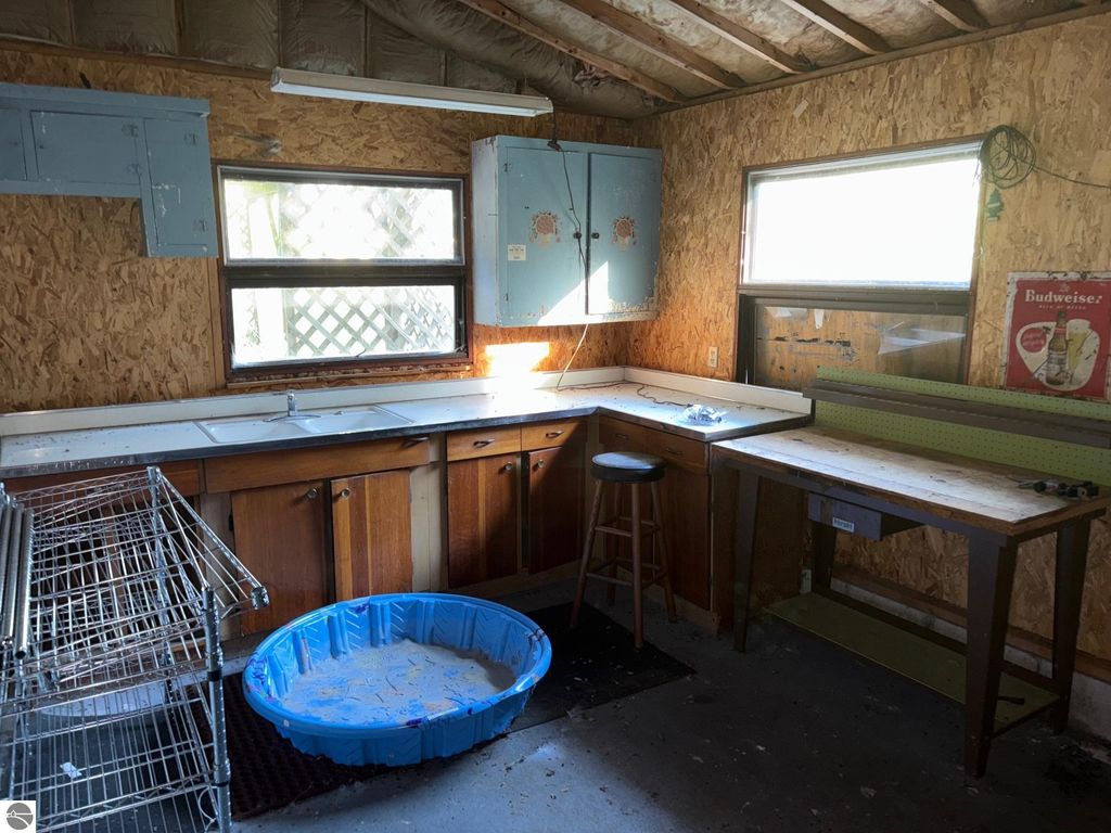 Interior of a workshop with wooden cabinets, a counter, a stool, and a blue plastic basin, featuring natural light from windows and a vintage Budweiser sign on the wall.