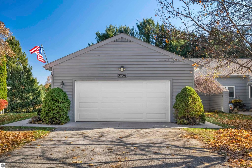 Condo exterior at 3756 Country Club Drive #9, Traverse City, MI, featuring a gray facade, white garage door, and American flag, surrounded by landscaped greenery and autumn leaves.