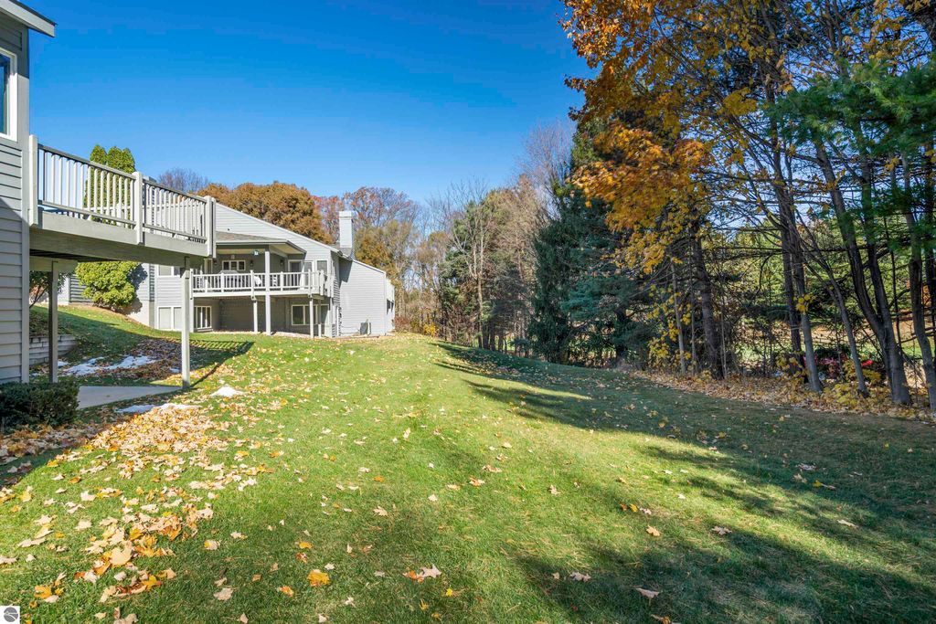 Exterior view of a light-filled condo in Traverse City, featuring a spacious lawn with autumn leaves, a balcony overlooking a peaceful setting, and mature trees in the background, highlighting the tranquil atmosphere of Country Club Woods community.