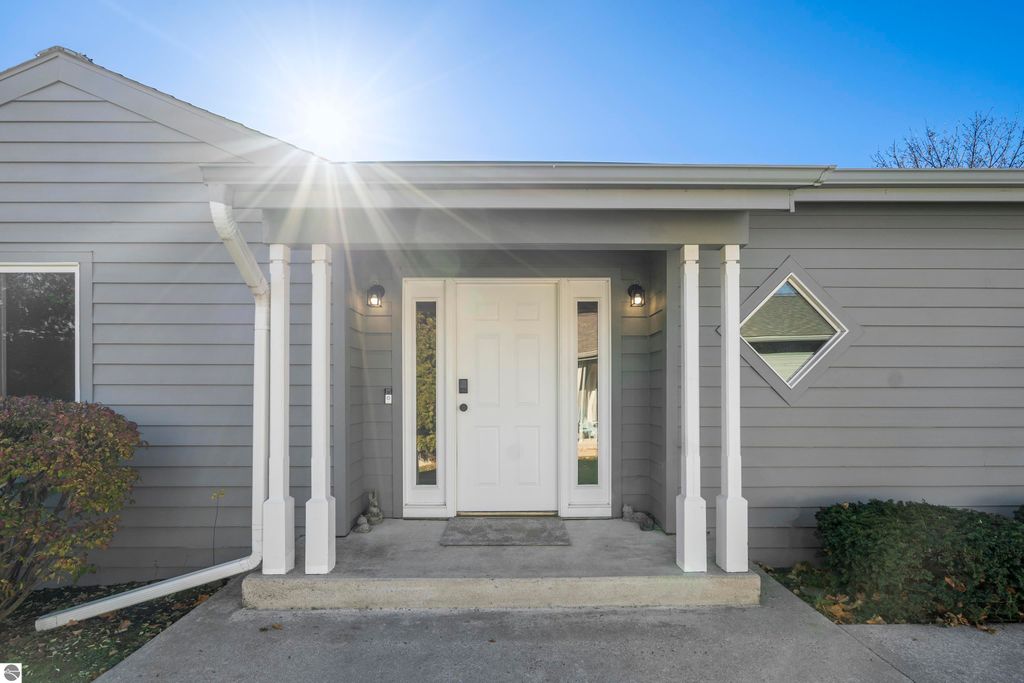 Front entrance of a modern 2-bedroom condo at 3756 Country Club Drive, Traverse City, featuring a white door, decorative columns, and a diamond-shaped window, surrounded by greenery and sunlight.