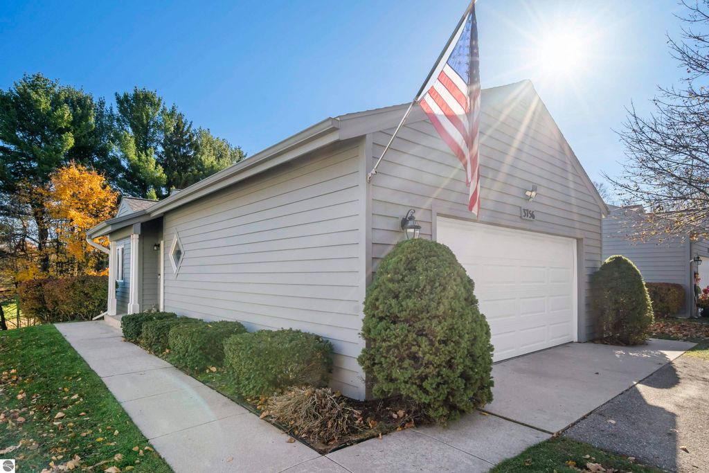 Exterior view of a 2-bedroom, 2-bath condo at 3756 Country Club Drive in Traverse City, featuring a well-maintained facade, American flag, landscaped yard, and garage, surrounded by trees and autumn foliage.