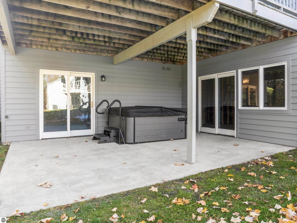 Patio area of a condo featuring a hot tub, sliding glass doors, and a concrete floor, surrounded by a grassy lawn with autumn leaves, located in the Country Club Woods community of Traverse City.