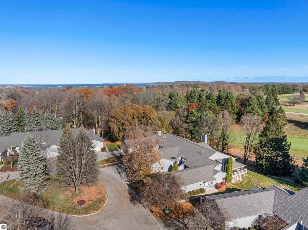 Aerial view of the Country Club Woods community in Traverse City, showcasing well-maintained condos, mature trees, and proximity to a golf course, with a clear blue sky and distant water views.