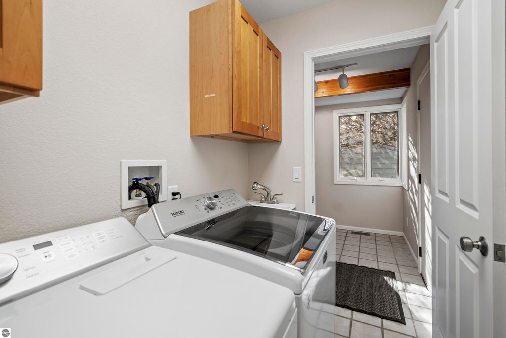 Washer and dryer in a laundry room with natural light from a window, featuring wooden cabinets and tiled flooring, part of a 2-bedroom, 2-bath condo for sale in Traverse City, MI.