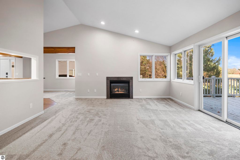Light-filled living room in updated Traverse City condo, featuring vaulted ceilings, cozy gas fireplace, large windows with scenic views, and access to a balcony overlooking mature trees and golf course.