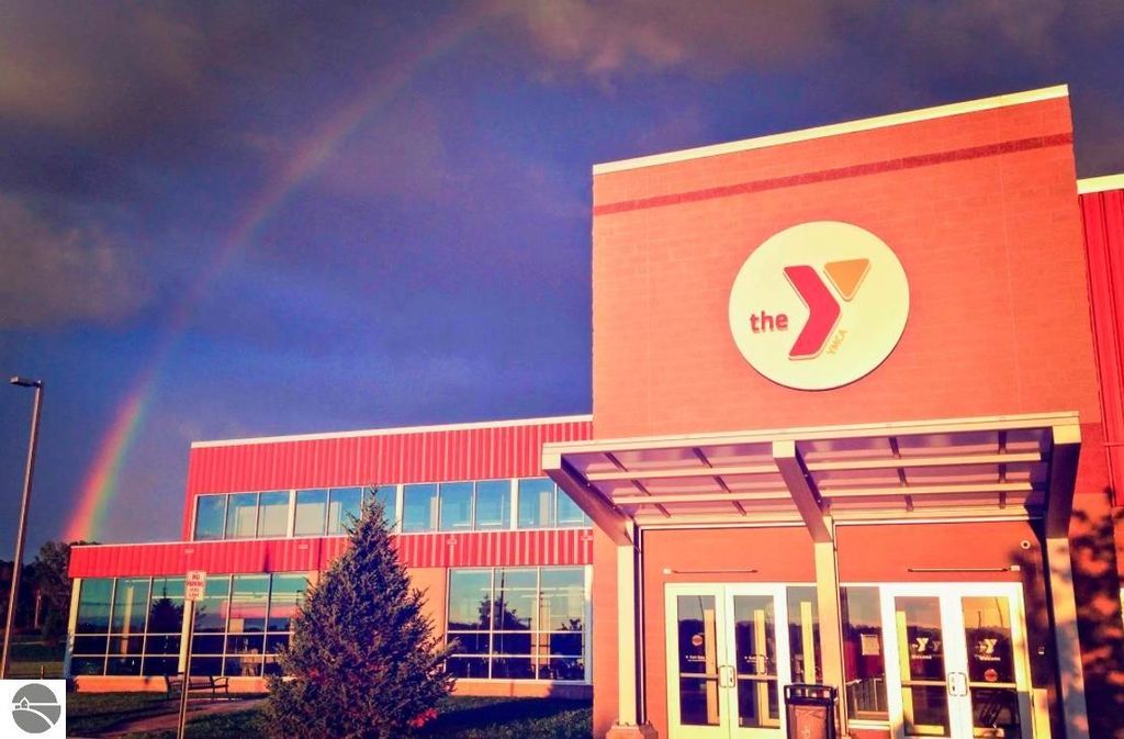 YMCA building exterior with logo, featuring large windows, a rainbow in the background, and surrounding greenery.