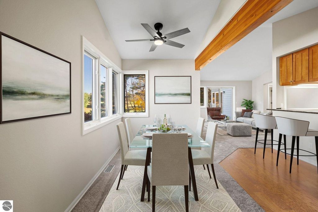 Dining area featuring a glass table set for four, surrounded by modern chairs, with large windows providing natural light and views of mature trees, complemented by contemporary artwork on the walls, in a stylish condo at 3756 Country Club Drive, Traverse City, MI.