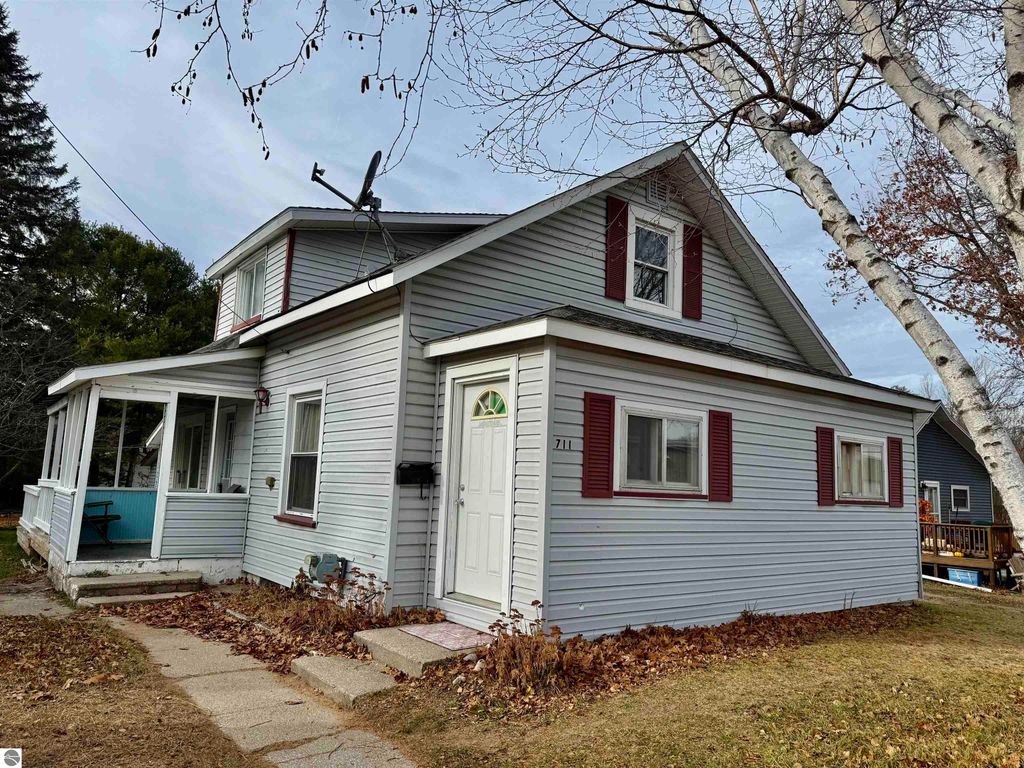 Exterior view of 711 Francisco Avenue, Alma, MI, featuring a two-story home with gray siding, red shutters, and a welcoming front porch, surrounded by a grassy yard and autumn foliage.