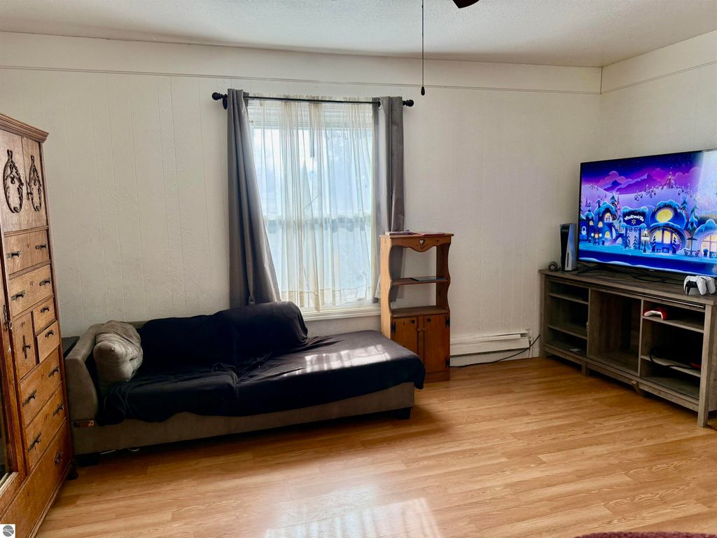 Living room interior featuring a couch, wooden dresser, and TV, showcasing a bright window with sheer curtains, emphasizing a cozy atmosphere in the Alma, MI property listing.
