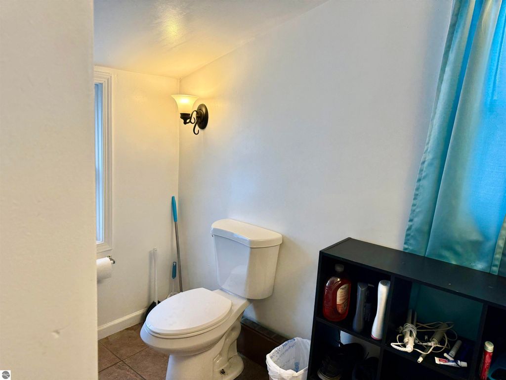 Bathroom interior featuring a white toilet, wall-mounted light fixture, and teal curtain, with a storage unit displaying personal care items.