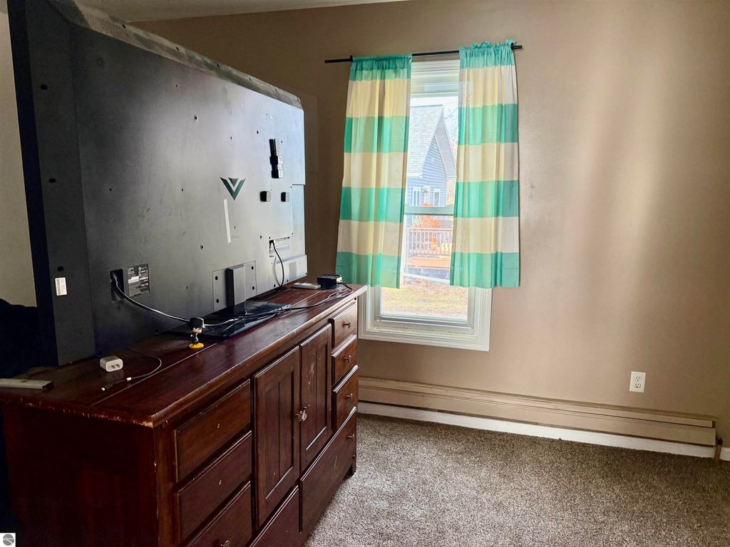 Interior view of a bedroom featuring a large television mounted on a dresser, striped curtains framing a window, and light brown walls, highlighting the room's spaciousness suitable for a rental property.