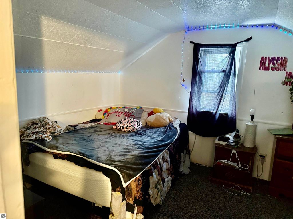 Cozy bedroom with a bed adorned with blankets and pillows, featuring a window with sheer curtains, decorative LED lights along the wall, and a bedside table with a lamp, showcasing a personal living space in a rental property.