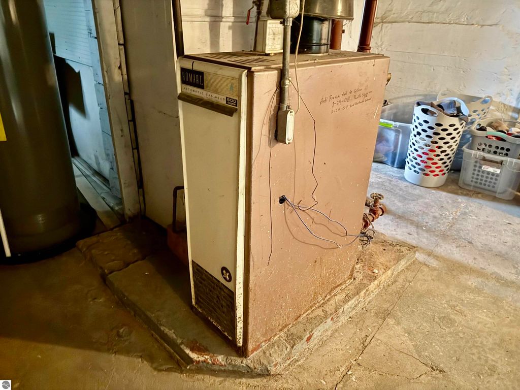 Basement heating unit, HOMART brand, with visible wires and plumbing, surrounded by storage bins and a water heater, relevant to property listing at 711 Francisco Avenue, Alma, MI.