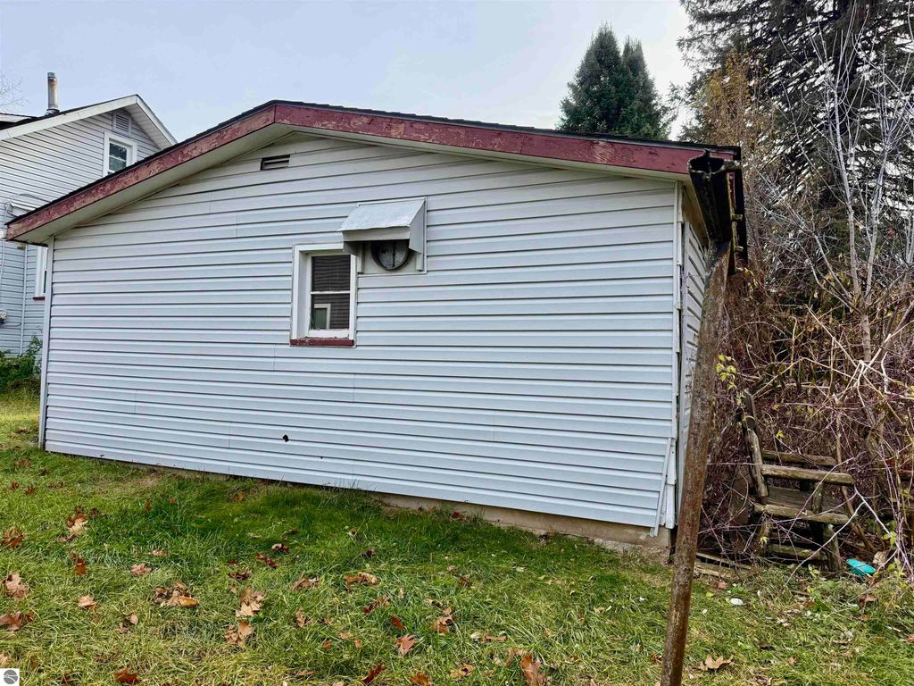 Exterior view of a single-story home at 711 Francisco Avenue, Alma, MI, featuring white siding, a red roof, and a side window, with overgrown grass and shrubs in the backyard.