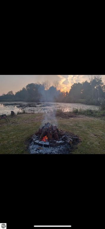 Campfire with smoke rising, surrounded by grassy area and pond, at sunset near 4419 10 Mile Road, Evart, MI, showcasing country living and outdoor space on the 14.5-acre property.
