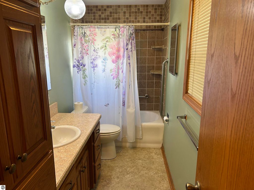 Bathroom featuring a floral shower curtain, beige countertop with sink, wooden cabinetry, and tiled walls, showcasing a clean and functional space in a home for sale at 11138 W 2nd Street, Fowler, MI.