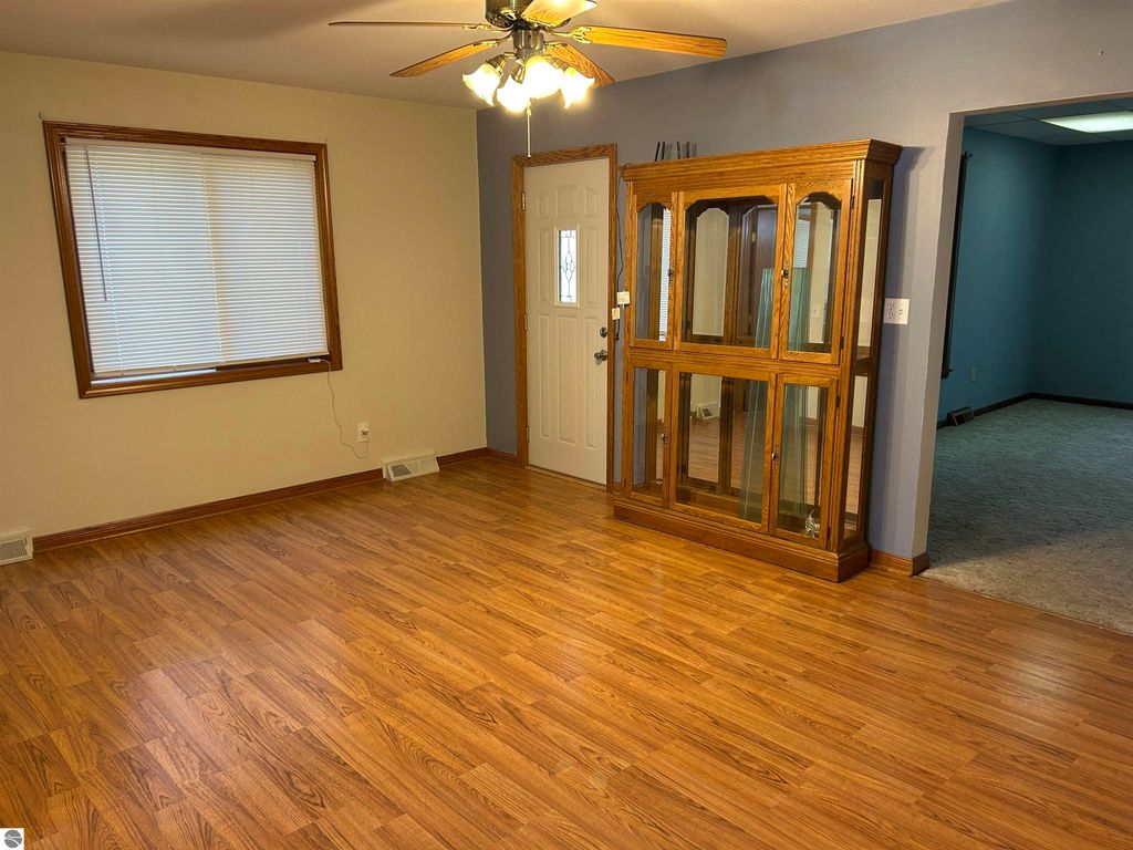 Living room interior of a home for sale in Fowler, MI, featuring hardwood floors, a ceiling fan, a large window with blinds, a wooden display cabinet, and a doorway leading to another room.