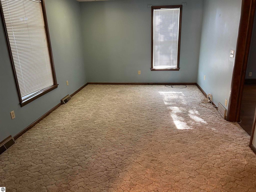 Empty living room with light blue walls, large windows with blinds, and beige carpet, showcasing potential for family gatherings in the Fowler, MI home listing.