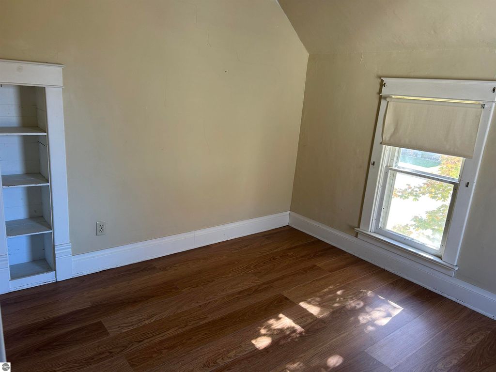 Cozy bedroom with hardwood flooring, window with blinds, and built-in shelves, showcasing potential for a personal space in a Fowler, MI home listing.