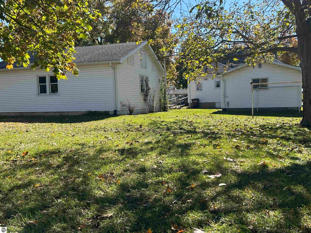 Backyard view of a 4-bedroom home at 11138 W 2nd Street, Fowler, MI, featuring two white houses, mature trees, and a grassy area with fallen leaves.