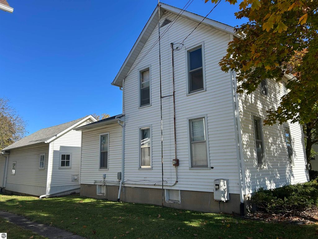 Exterior view of a two-story white house at 11138 W 2nd Street, Fowler, MI, featuring multiple windows, a side garage, and surrounding greenery, highlighting its proximity to downtown amenities.